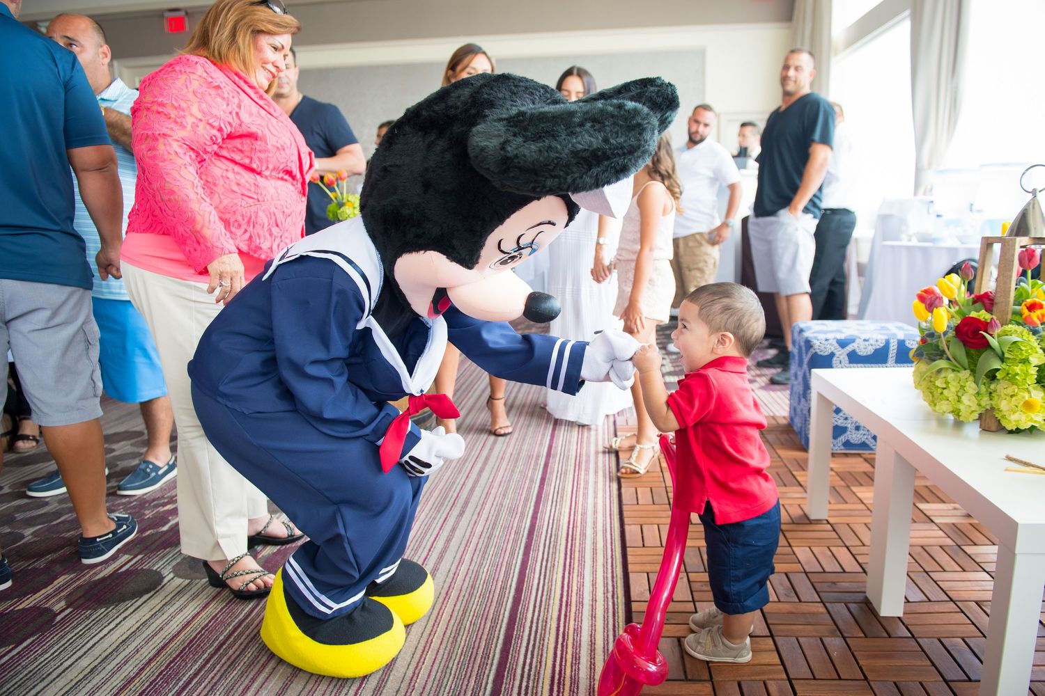 A Minnie Mouse in a sailor outfit bends down to interact with a small child wearing red on a covered porch.