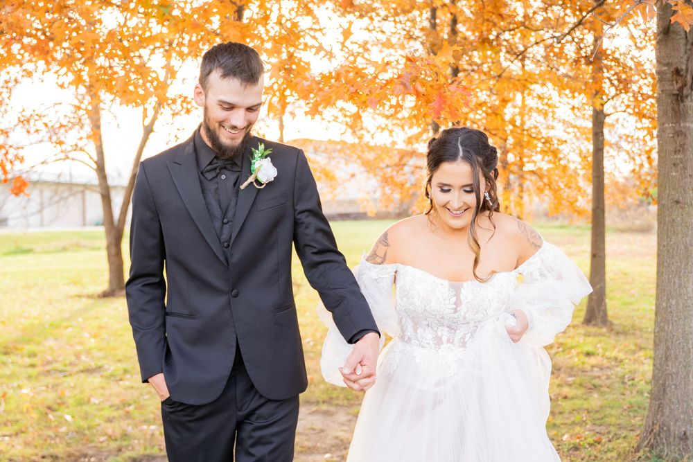 Smiling bride and groom holding hands walking together beneath vibrant orange fall trees.