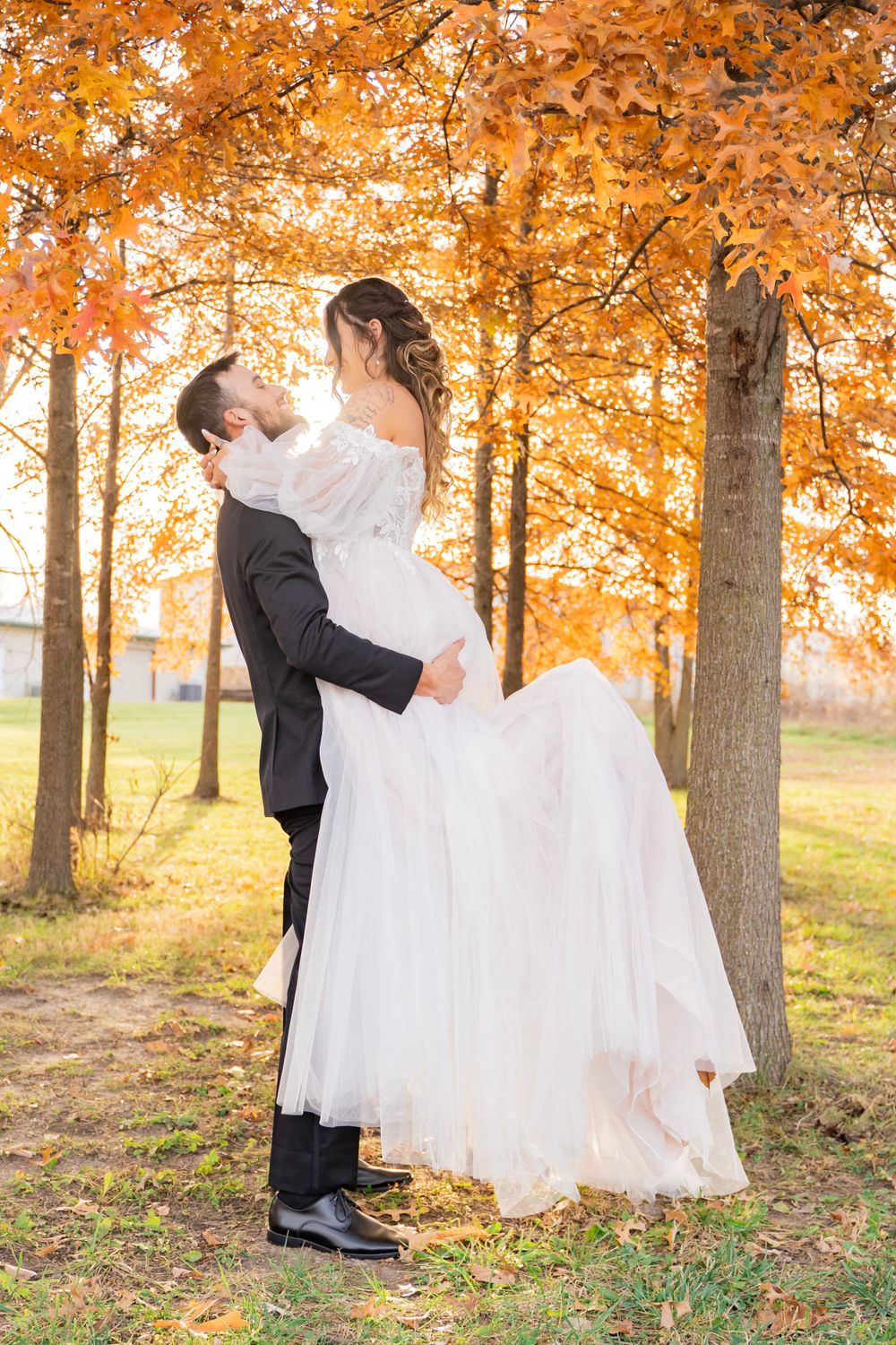 Groom lifting bride in white tulle gown for a kiss under glowing orange fall foliage.