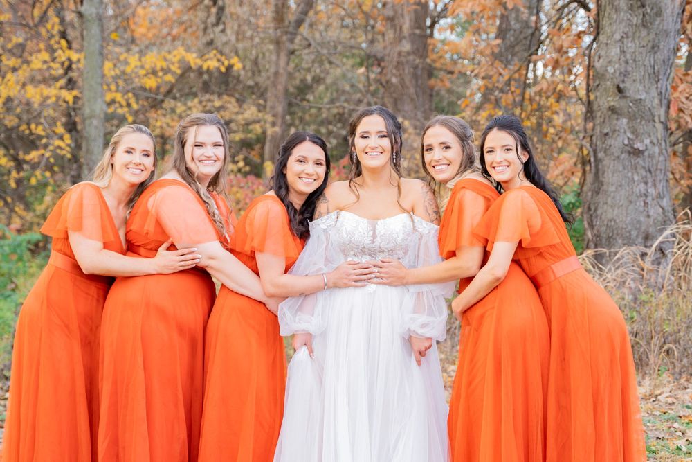 Bride in white gown poses with five bridesmaids in orange dresses in autumn woodland setting.