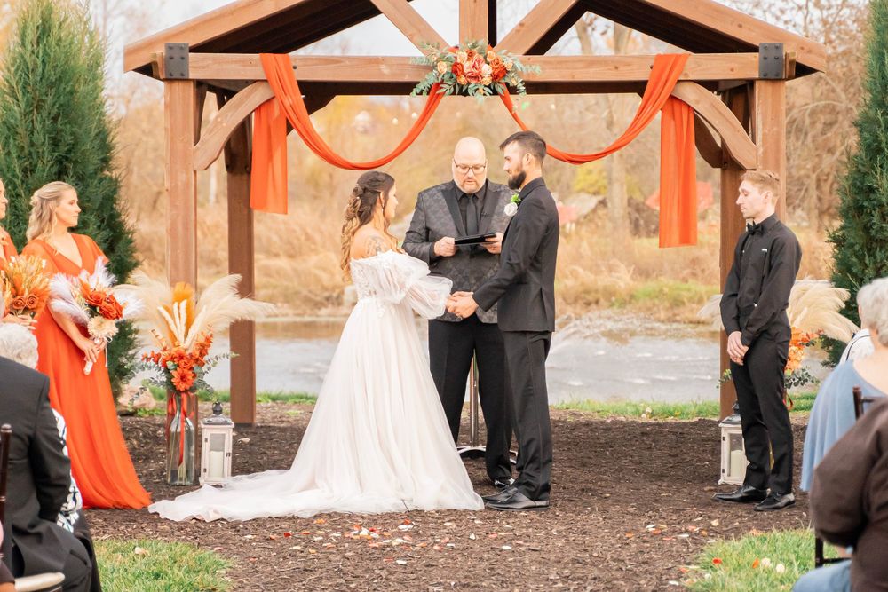 Bride and groom exchange vows under a wooden arch with orange floral decor at outdoor ceremony.