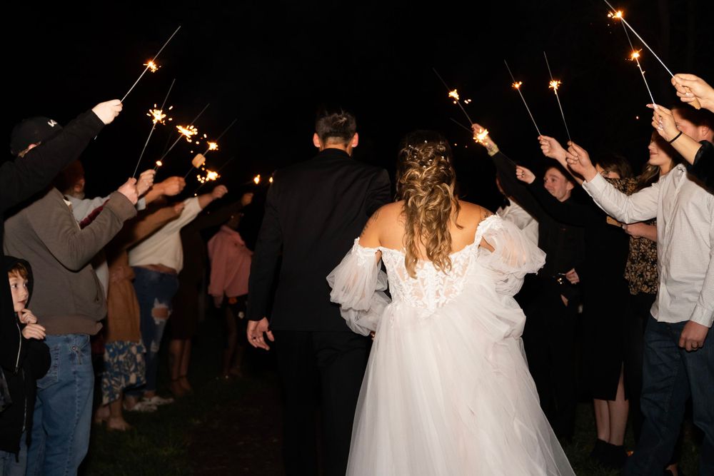 Bride and groom walking through sparkler send-off tunnel with wedding guests at night.