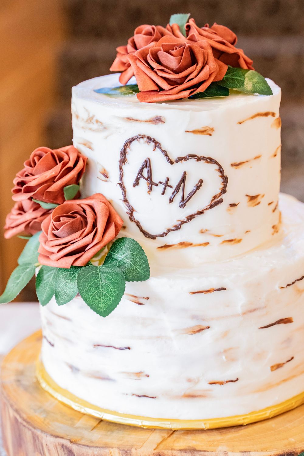 Two-tier white birch bark wedding cake with rust roses and carved initials A+N in a heart design.