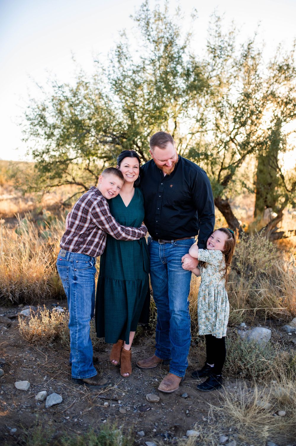 Family of four hugging and laughing outdoors in a desert setting during golden hour.