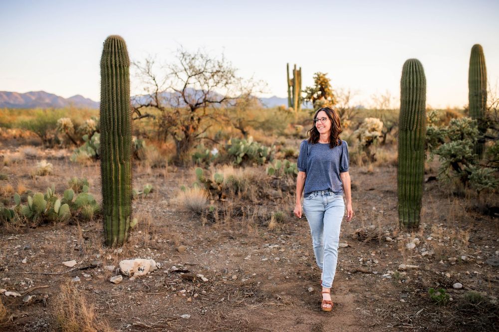Young woman walking between tall saguaro cacti on desert trail with mountains in background.