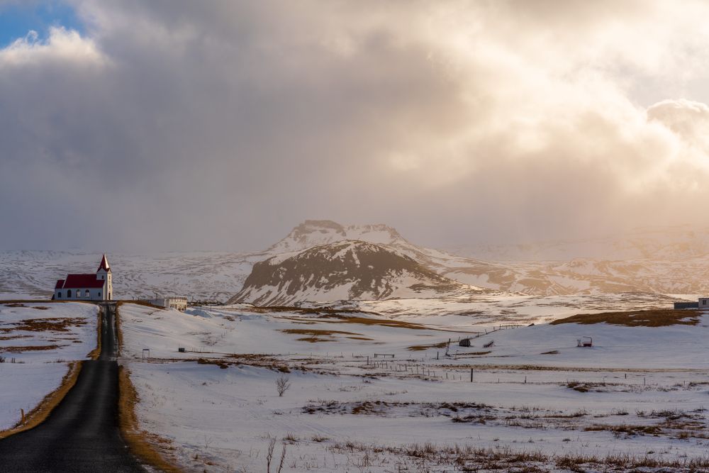 A long rural road leads to a picturesque church next to a wintery Icelandic mountain range and a dramatic backlit cloudy sky.