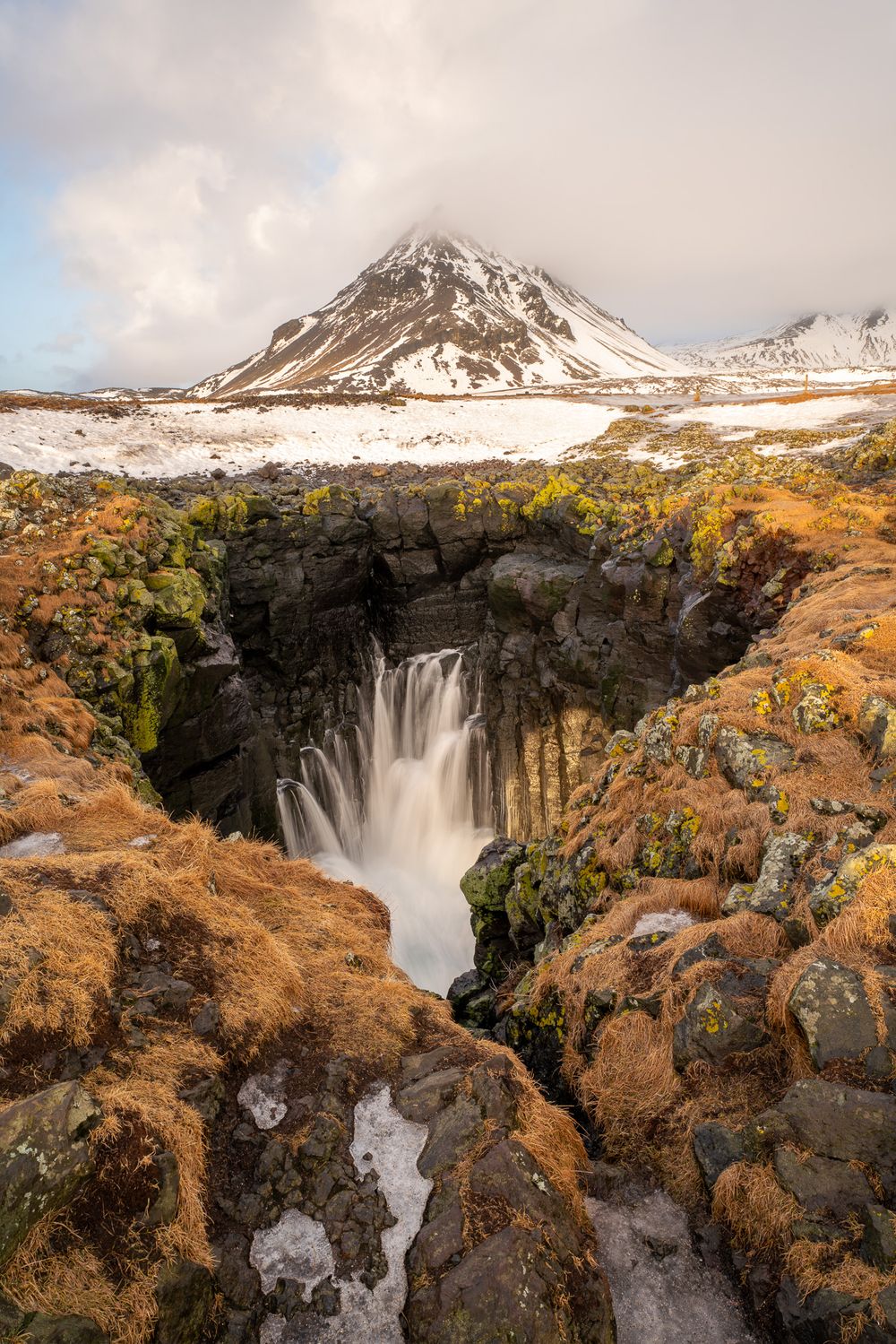 A basalt cliff cave along the Icelandic coast creates the illusion of a waterfall in front of a snowy mountain peak shrouded by clouds
