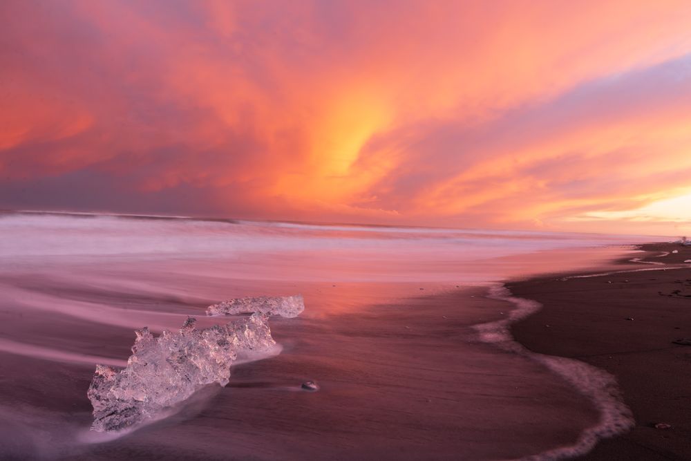 A crystalline chunk of ice sits on the black sand beach of Jokulsarlon, aka Diamond Beach beneath a dynamic pink orange sunset sky.