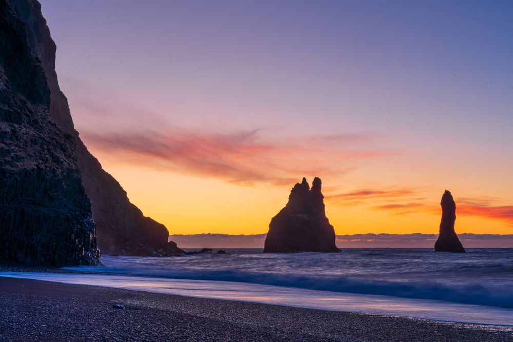 Black basalt sea stacks rise from the ocean along Iceland's southern shore at dawn. A gold, pink and indigo sky creates the background.