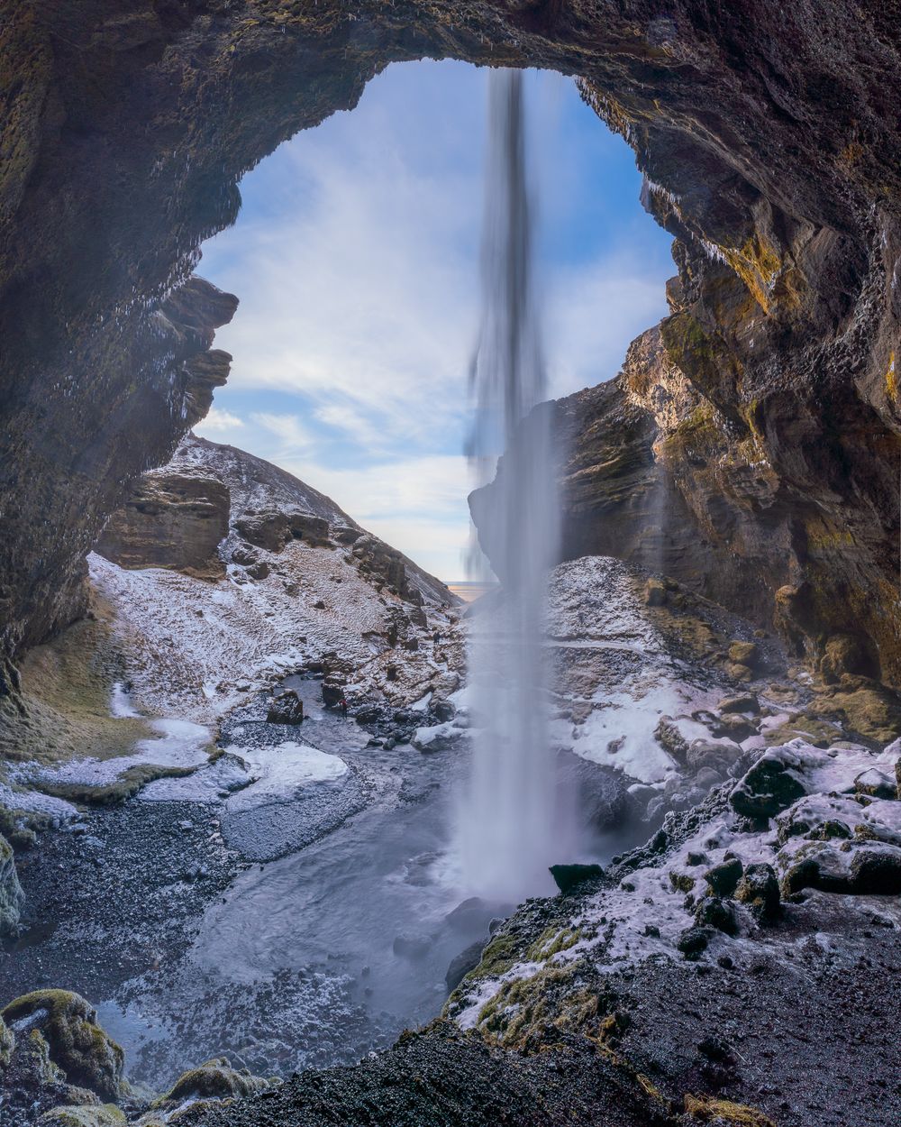Behind Kvernufoss. A waterfall streams down from an arch-like opening to a rocky pool below. The falls are surrounded by a rocky cavern.