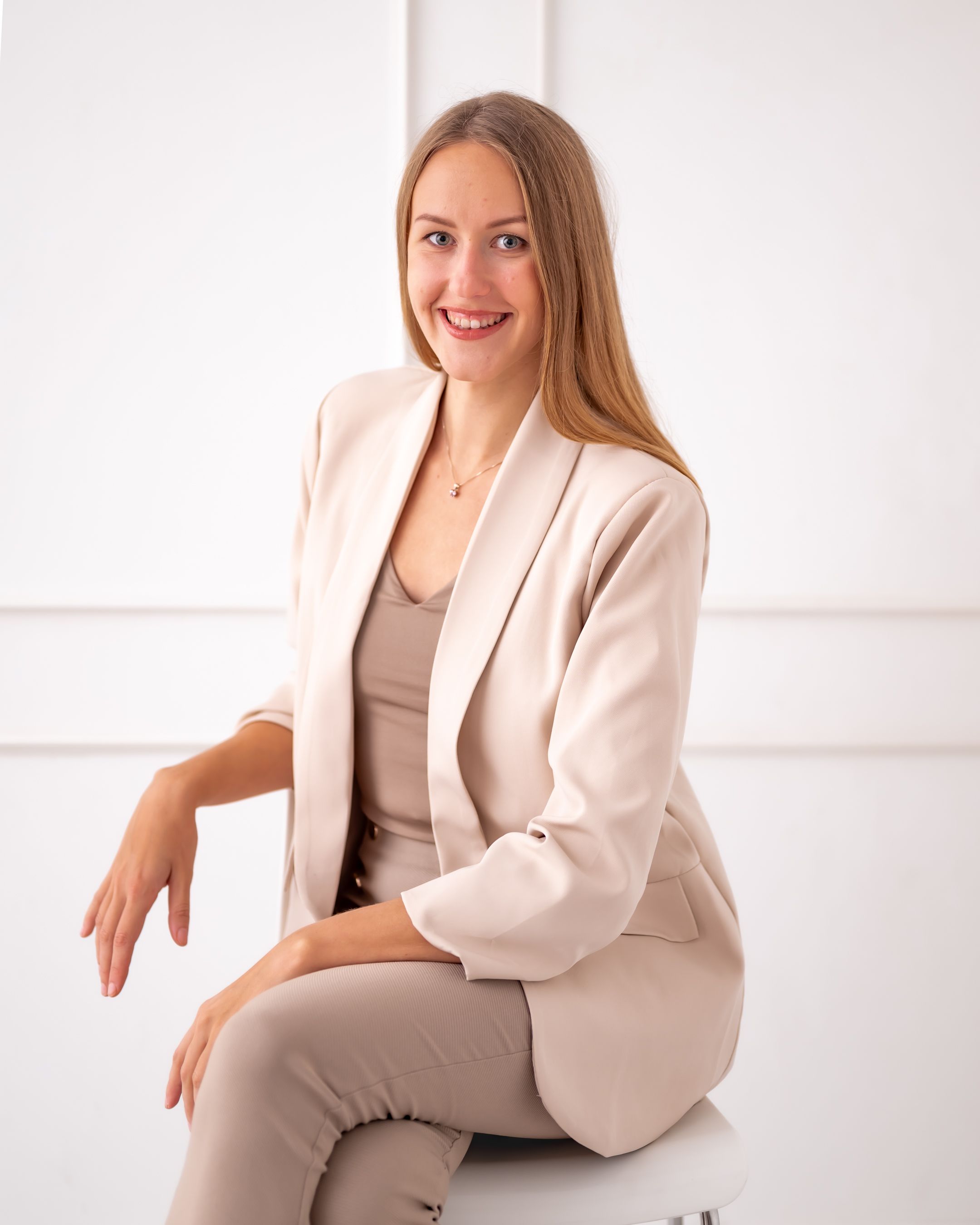 Businesswoman wearing cream colored suit sits elegantly on white chair in minimalist setting.