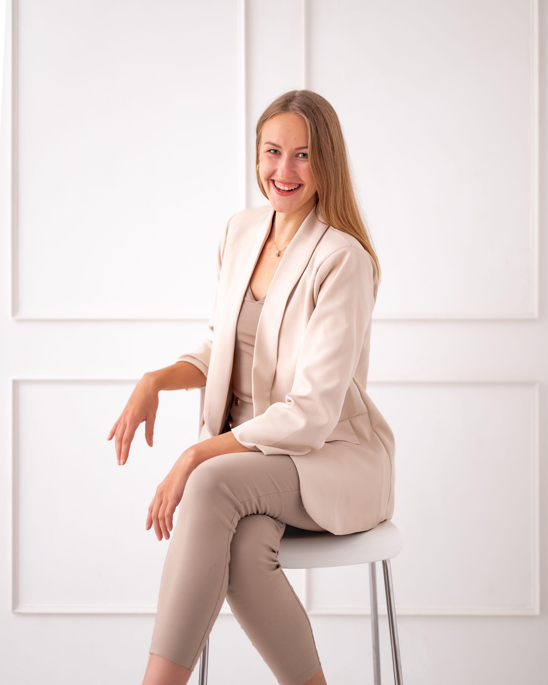 Professional woman in beige blazer and pants sits poised on chair against white wall.