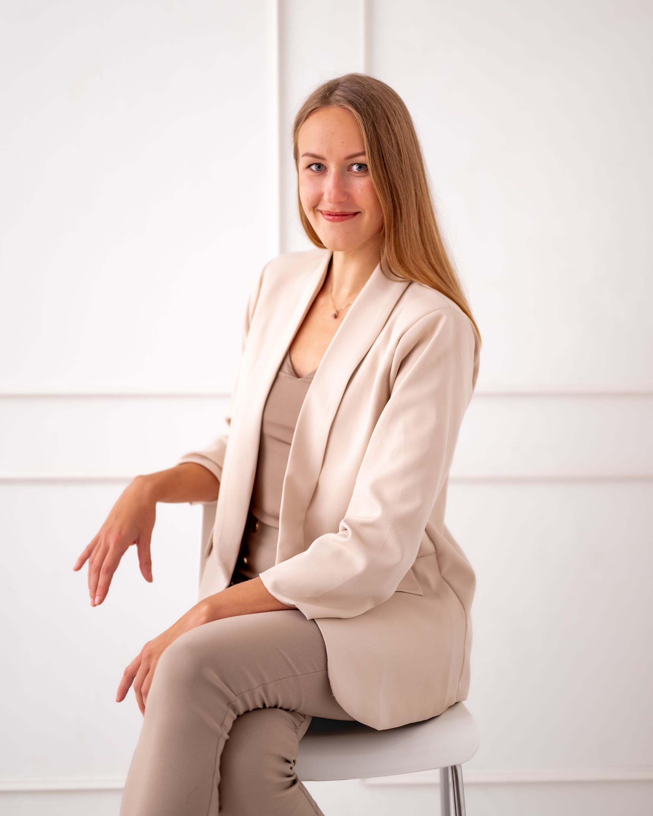 Professional portrait in neutral tones featuring seated pose against white paneled wall.