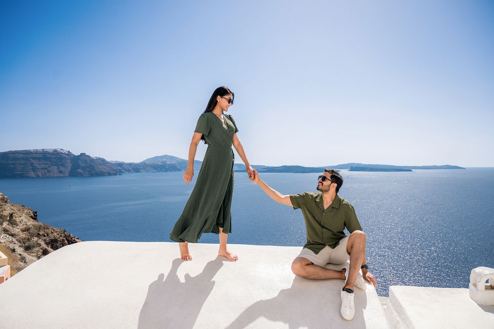 A couple stands on a white rooftop terrace overlooking the beautiful blue sea and mountains of Santorini, Greece.
