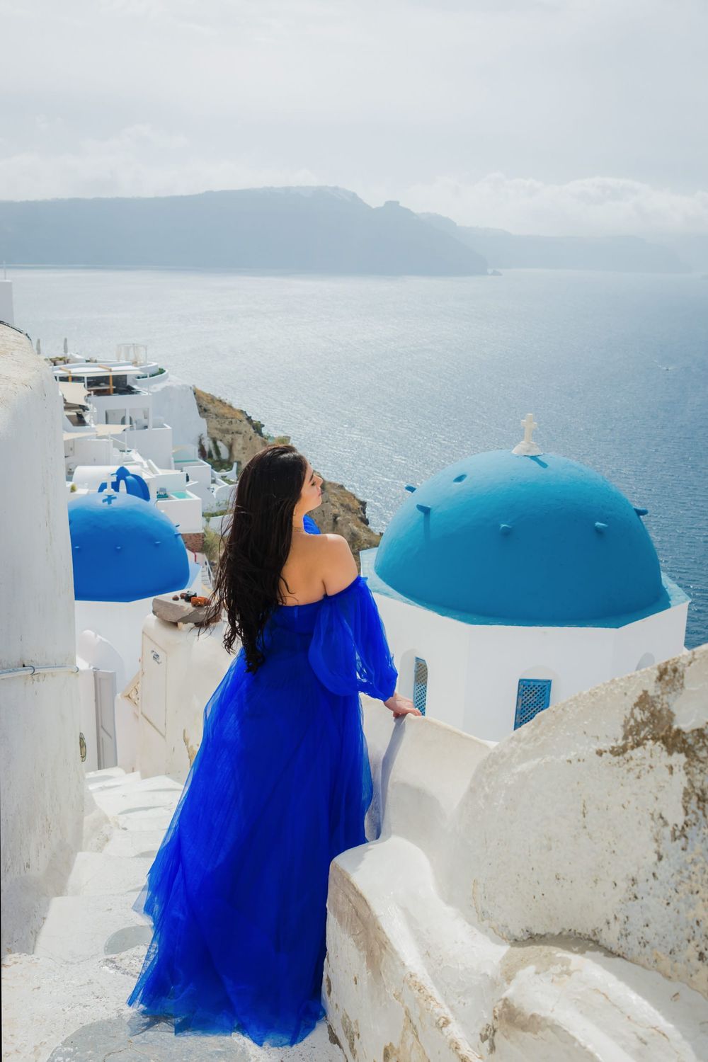 Person in flowing blue dress gazes at iconic white buildings and blue domes of Santorini, Greece against ocean view.
