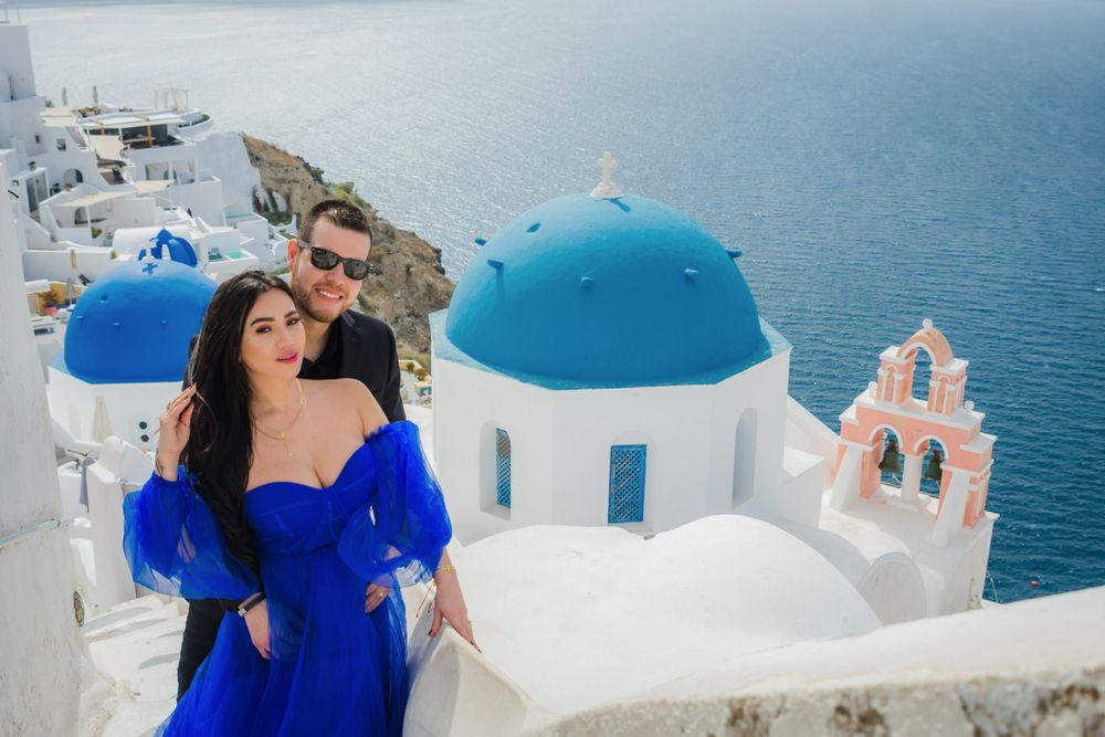 Tourist in blue dress poses against iconic white buildings and blue domed church in Santorini, Greece.