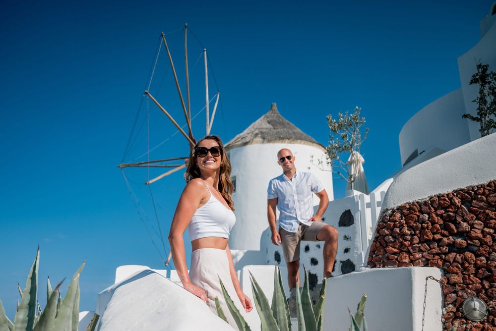Two people pose near a traditional white windmill in Santorini, Greece against a bright blue sky.