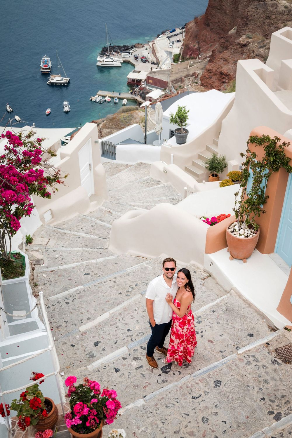 Stone steps lead down to the Mediterranean Sea in Santorini, Greece, with bright pink bougainvillea flowers and white buildings.