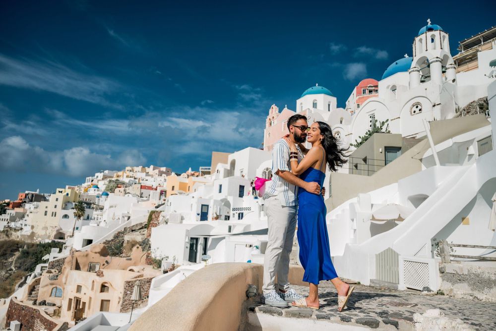 A traveler in a blue dress stands on a pathway in Santorini, Greece with white buildings and blue domes in the background.