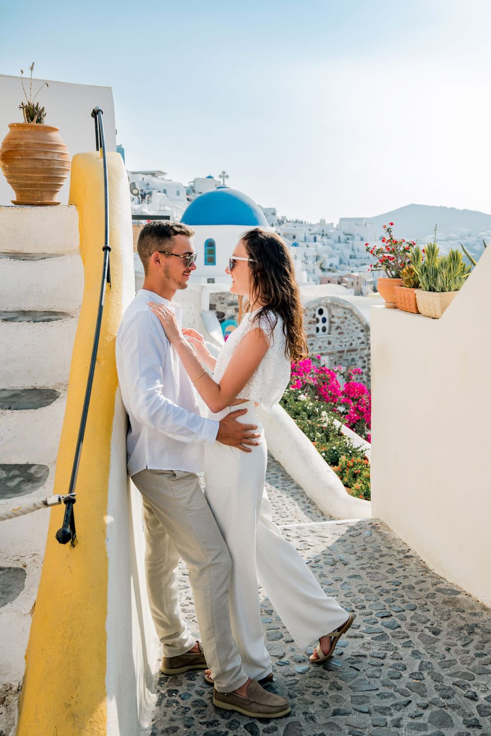 A romantic couple embraces on a charming cobblestone path in Santorini, Greece with iconic blue domes and white buildings.
