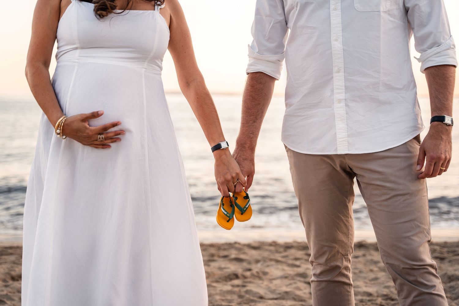 Pregnant couple holding hands and tiny baby sandals on the beach