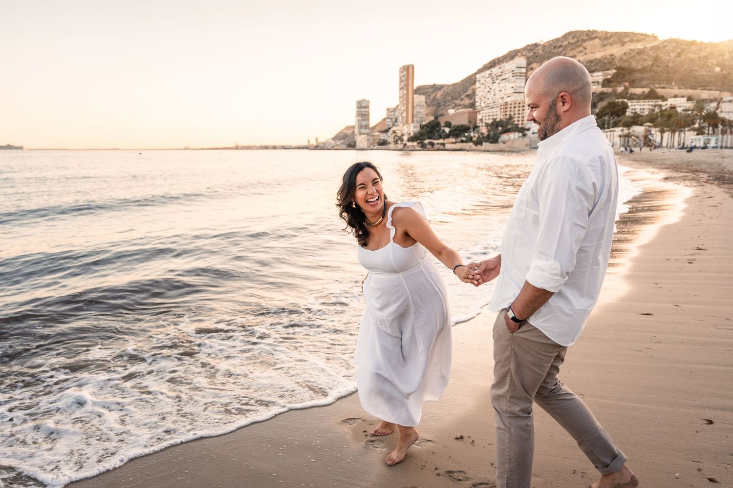 Couple smiling and laughing and holding hands on the beach at sunset with golden sand and glow of sun