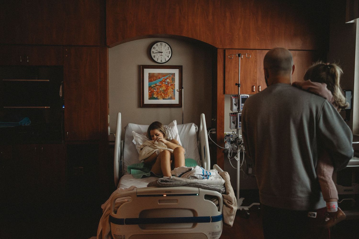 Family gathers around hospital bed in warmly lit medical room with wooden cabinets.