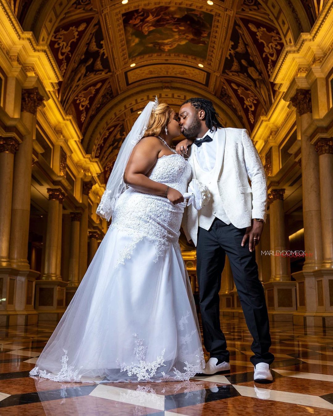 Newlyweds kissing in an ornate grand hall, bride in mermaid gown, groom in white tuxedo.