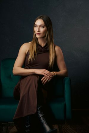 Woman in brown sleeveless dress and black boots seated in teal velvet chair against dark background.