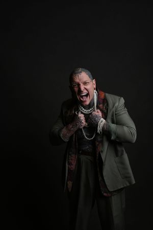 Tattooed man in olive suit screaming while holding pearl necklace against black background.