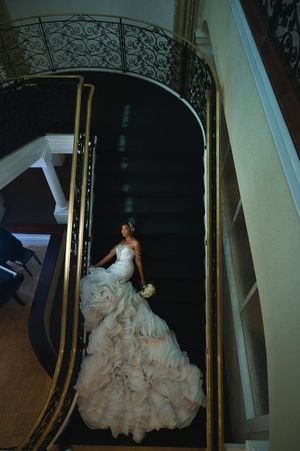 Bride in a stunning ruffled blush wedding gown descending an ornate curved staircase with gold railings.