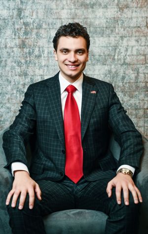 Young professional man in dark plaid suit with red tie smiling confidently in studio portrait.