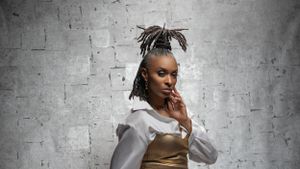 Stylish Black woman with locs updo in gold crop top and white shirt against white brick wall.
