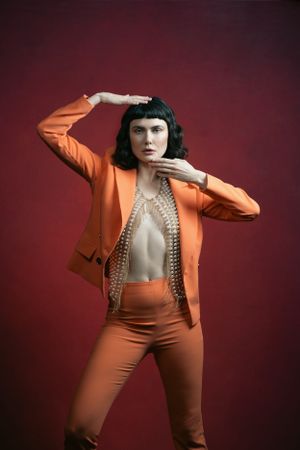 Bold fashion model in orange suit posing dramatically against deep red studio background.