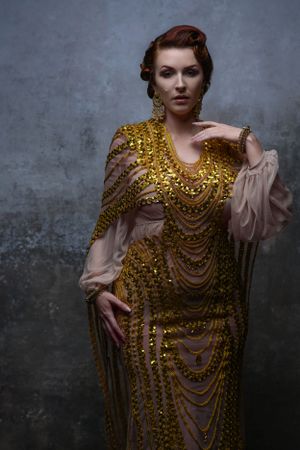 Elegant woman in ornate gold beaded gown with sheer sleeves posing against gray studio backdrop.