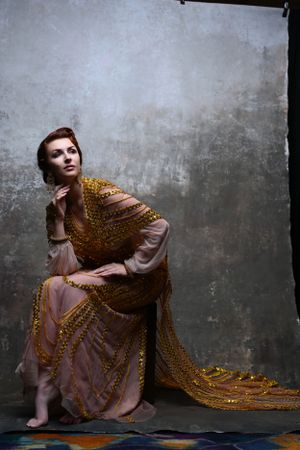 Graceful woman in gold embellished pink gown seated on floor against gray textured studio wall.