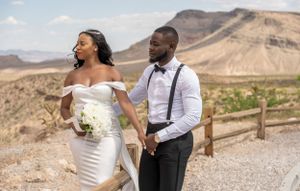 Bride and groom pose at desert overlook, she in white off-shoulder gown holding bouquet, he in suspenders.