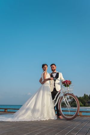 Bride in ball gown and groom in tuxedo pose with vintage bicycle on waterfront dock at sunset.
