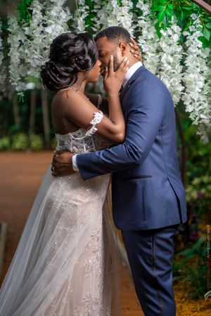 Bride and groom share first kiss under white floral arch in elegant indoor wedding ceremony.