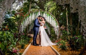 Couple shares romantic kiss under floral arch in lush tropical garden wedding ceremony setting.