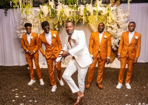Groom in white suit dances with four groomsmen in matching orange suits at wedding reception.