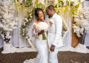Bride and groom smile together before white floral backdrop, both dressed in elegant white wedding attire.