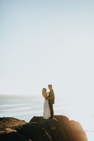 Bride and groom embrace on rocky coastal cliff at sunset with ocean horizon in the background.