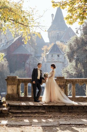 Bride in blush gown and groom in black suit pose on stone terrace with autumn castle backdrop.