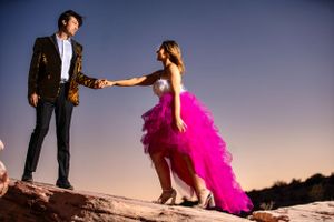 Couple in formal attire holding hands on rocky terrain at dusk, woman in pink tulle gown.