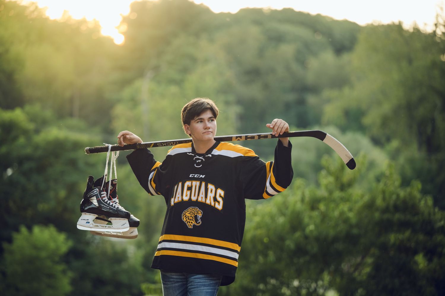 Teen in a Jaguars hockey jersey holding a stick and skates over his shoulders in golden sunlight.