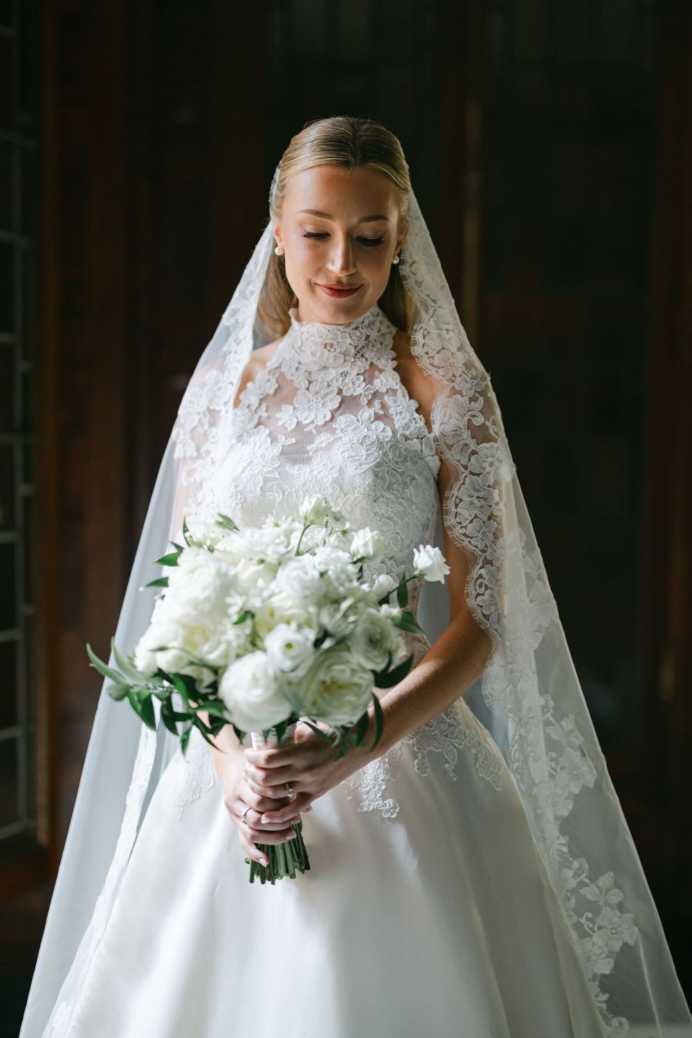 Elegant bride in lace wedding gown and veil holding white floral bouquet of peonies indoors. - The Blackstone Hotel Wedding