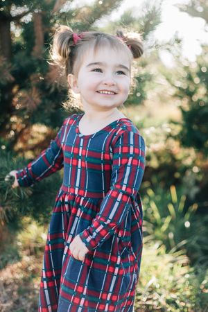 Child in red and blue plaid dress smiling in a sunny garden setting.