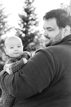 Black and white portrait of father holding baby against evergreen trees.