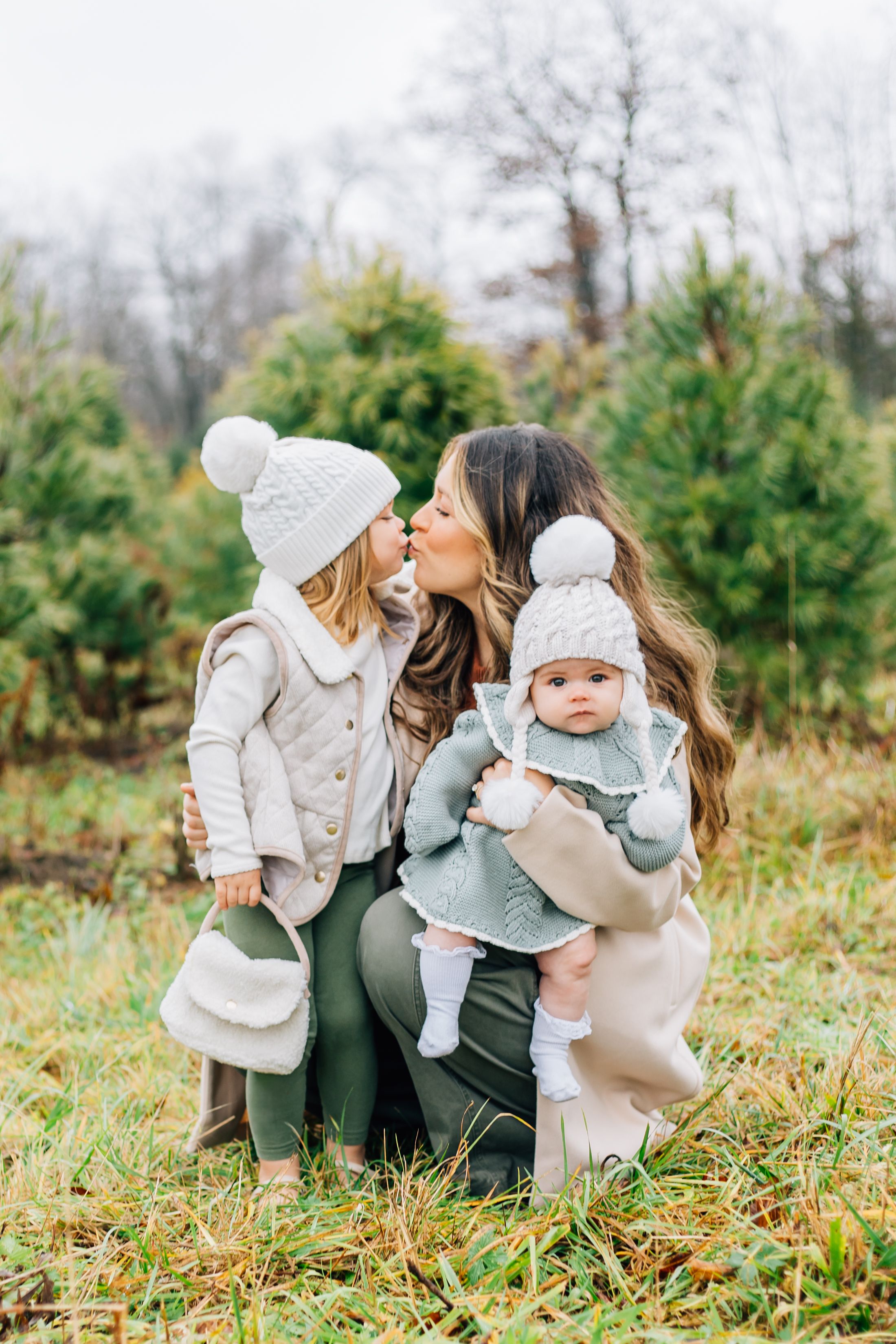 A cozy winter family portrait among evergreen trees with matching white knit hats and neutral colored outfits.